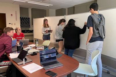 Four PLTL students are drawing components of a foodweb on the board while two students provide support and feedback while seated at a table.