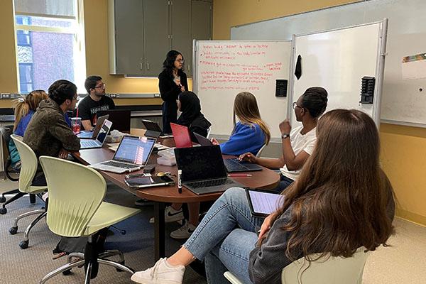 A PLTL peer leader collects verbal responses and writes them on a white board. Seven students sit around a table participating in the discussion.